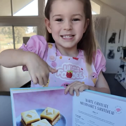 Child in a pink apron holding a book about making 'Millionaire's bars' that are described as 'but toddler-friendly'.